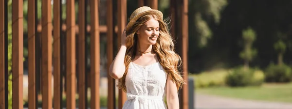 Plano panorámico de hermosa chica en vestido blanco tocando sombrero de paja y sonriendo con los ojos cerrados - foto de stock