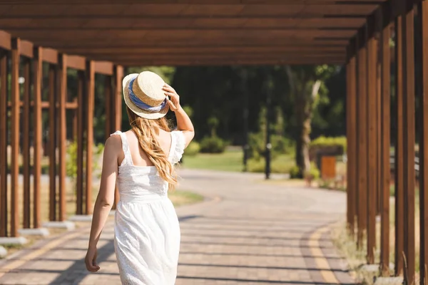 Vue arrière de la fille en robe blanche touchant chapeau de paille tout en marchant sur le chemin près de la construction en bois — Photo de stock