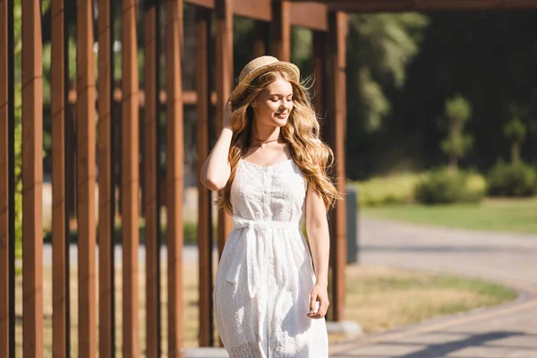 Beautiful girl in white dress touching straw hat and smiling with closed eyes — Stock Photo