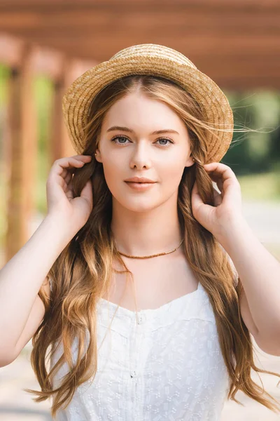 Retrato de la foto de la hermosa chica en sombrero de paja tocando el pelo mientras mira a la cámara - foto de stock