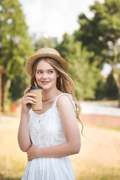 Hermosa chica en vestido blanco y sombrero de paja sosteniendo taza de café de papel, sonriendo y mirando hacia otro lado - foto de stock