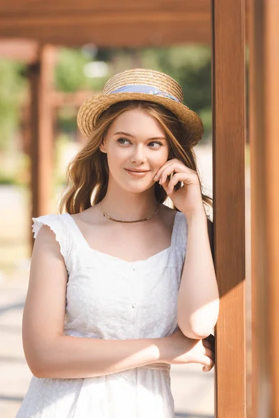 Hermosa chica en vestido blanco y sombrero de paja apoyado en la construcción de madera - foto de stock