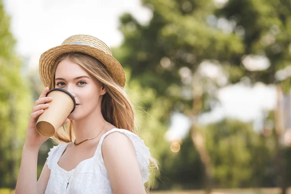 Bella ragazza in abito bianco e cappello di paglia in possesso di tazza di caffè di carta e guardando la fotocamera — Foto stock