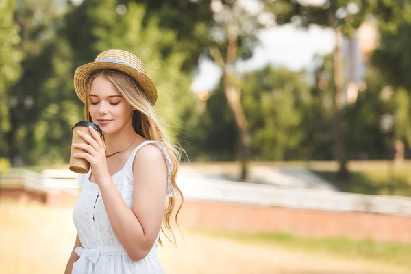 Hermosa chica en vestido blanco y sombrero de paja sosteniendo taza de papel y beber café con los ojos cerrados - foto de stock