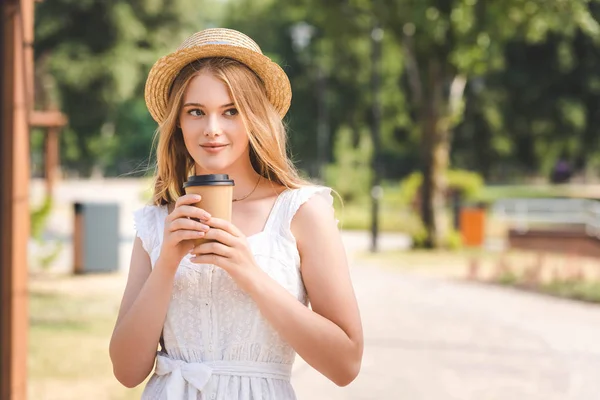 Schönes Mädchen in weißem Kleid und Strohhut hält Pappbecher in der Hand und schaut weg — Stockfoto