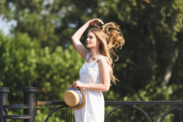 Красивая девушка в белом платье, держащая соломенную шляпу, прикасаясь к волосам и отводя взгляд — стоковое фото
