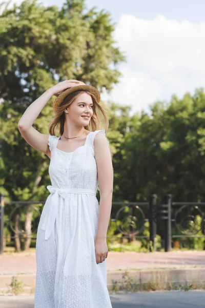 Hermosa joven en vestido blanco tocando sombrero de paja mientras sonríe y mirando hacia otro lado - foto de stock