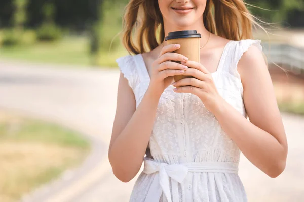 Vista recortada de chica joven en vestido blanco y sombrero de paja sonriendo y sosteniendo taza de café de papel - foto de stock
