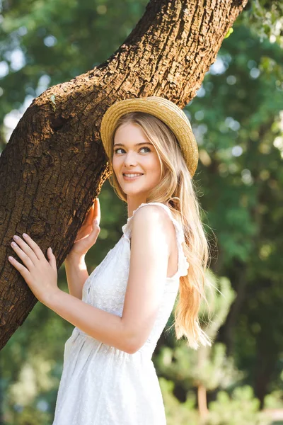Beautiful girl in white dress and straw hat standing near trunk of tree — Stock Photo