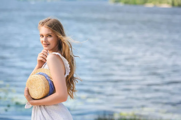 Hermosa joven de pie en la orilla del río mientras sostiene sombrero de paja y mirando a la cámara - foto de stock