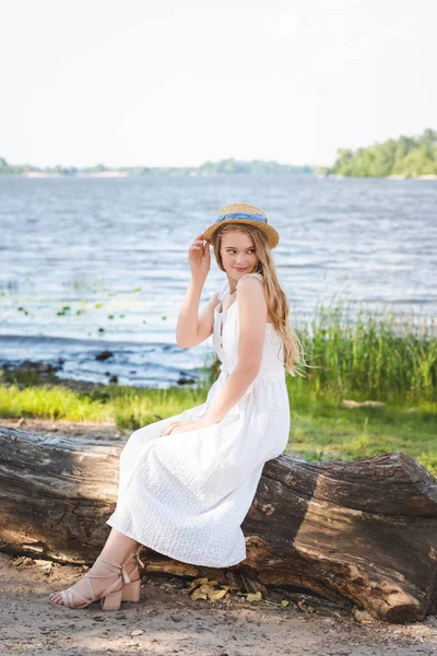 Belle fille en robe blanche et chapeau de paille assis sur le tronc de l'arbre sur la rive de la rivière et regardant loin — Photo de stock