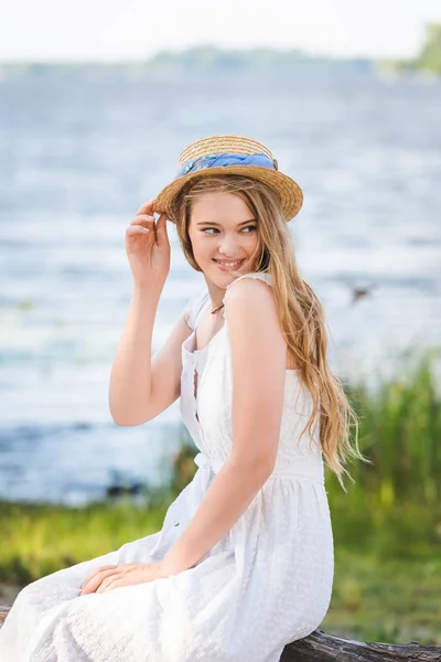 Menina bonita em vestido branco tocando chapéu de palha enquanto sentado no tronco da árvore na margem do rio e olhando para longe — Fotografia de Stock