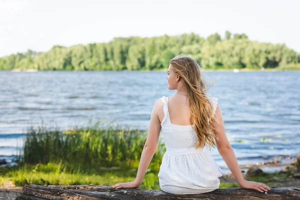 Vue arrière de la fille assise sur le tronc d'arbre près de la rive de la rivière et regardant loin — Photo de stock