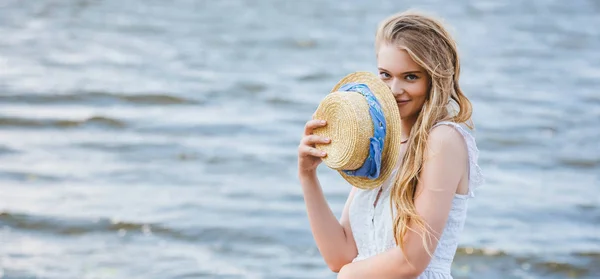 Panoramic shot of beautiful girl holding straw hat near face, smiling and looking at camera — Stock Photo