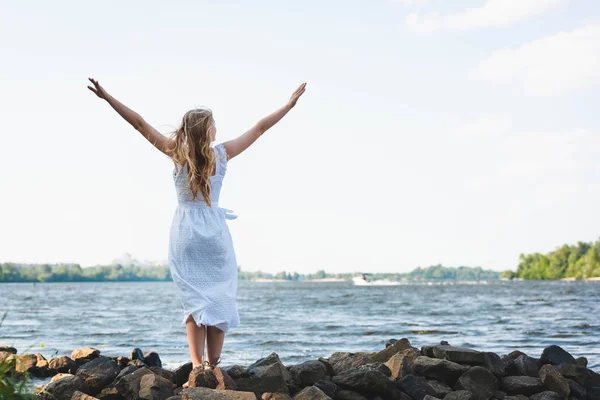 Повний вигляд дівчини в білій сукні, що стоїть на скелястому березі річки з руками в повітрі — стокове фото