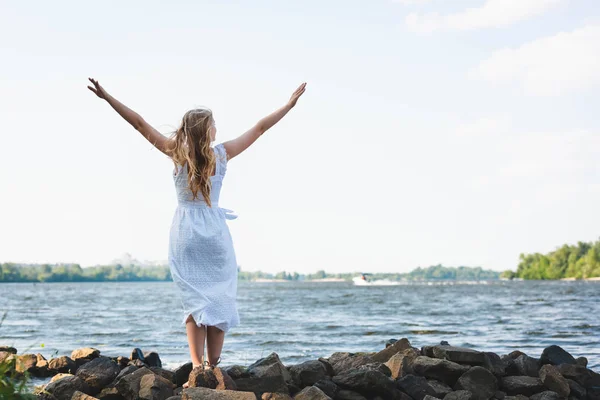 Vue pleine longueur de la fille en robe blanche debout sur le rivage de la rivière rocheuse avec les mains dans l'air — Photo de stock