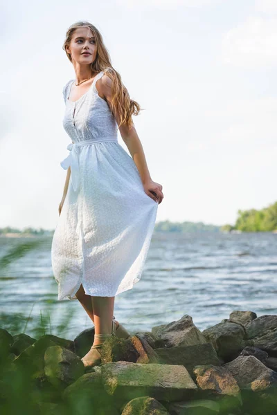 Selective focus of beautiful girl in white dress walking on rocky river shore and looking away — Stock Photo