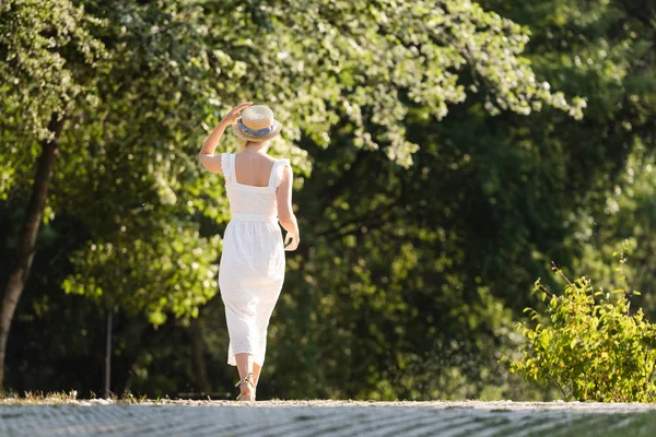 Back view of girl in white dress touching straw hat while walking in park — Stock Photo