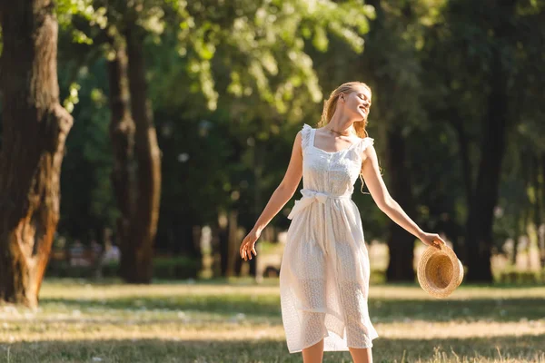 Menina bonita em vestido branco segurando chapéu de palha enquanto sorrindo e de pé no prado com os olhos fechados — Fotografia de Stock