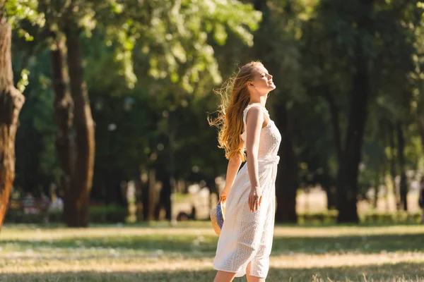Vista lateral da menina bonita em vestido branco segurando chapéu de palha enquanto sorrindo e de pé no prado com os olhos fechados — Fotografia de Stock