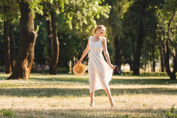 Vista completa de la hermosa chica en vestido blanco sosteniendo sombrero de paja mientras sonríe y de pie en el prado con los ojos cerrados - foto de stock