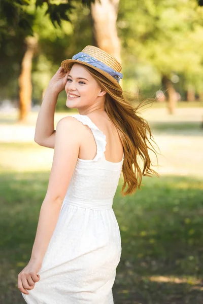 Hermosa chica en vestido blanco tocando sombrero de paja mientras y de pie en el prado, sonriendo y mirando hacia otro lado - foto de stock