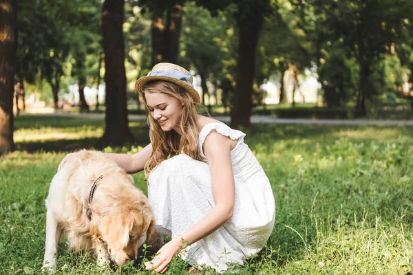 Beautiful girl in white dress and straw hat petting golden retriever while sitting on meadow and looking at dog — Stock Photo