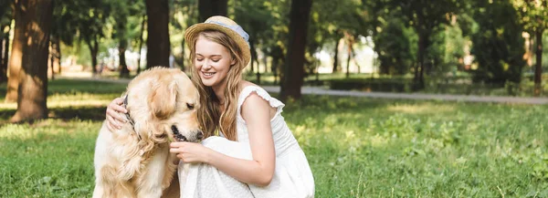 Panoramic shot of beautiful girl in white dress and straw hat petting golden retriever while sitting on meadow — Stock Photo