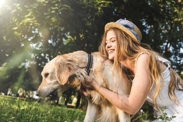 Hermosa joven en vestido blanco y sombrero de paja abrazando golden retriever mientras sonríe, sentado en el prado y mirando al perro - foto de stock