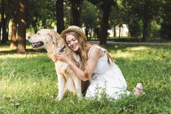 Beautiful young girl in white dress and straw hat hugging golden retriever while smiling and sitting on meadow with closed eyes — Stock Photo