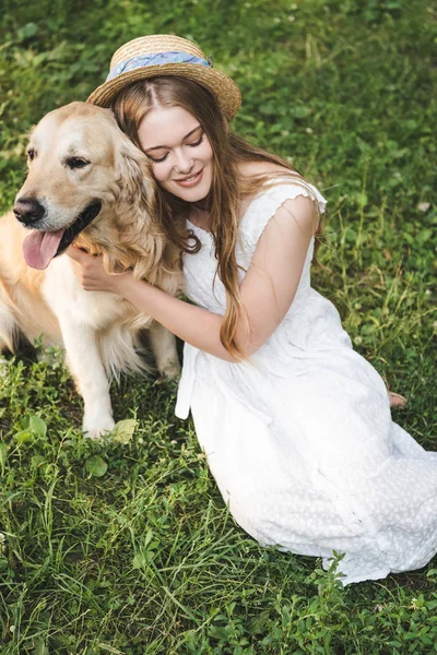 High angle view of beautiful young girl in white dress and straw hat hugging golden retriever while smiling and sitting on meadow with closed eyes — Stock Photo