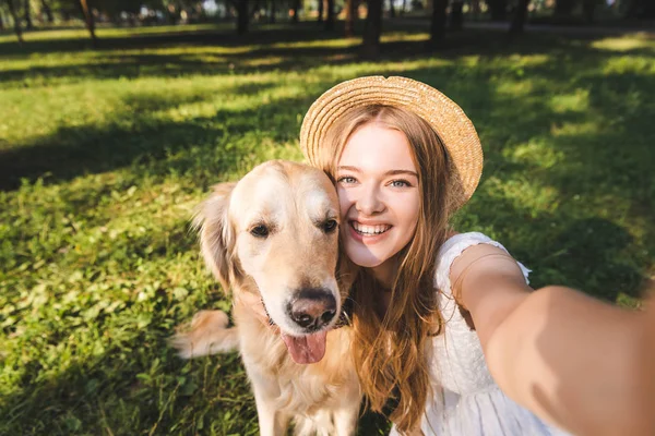 Beautiful young girl in white dress and straw hat hugging golden retriever and taking selfie while sitting on meadow, smiling and looking at camera — Stock Photo