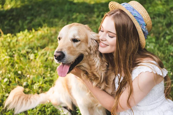 Hermosa chica en vestido blanco y sombrero de paja abrazando golden retriever mientras está sentado en el prado y mirando al perro - foto de stock