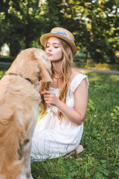 Hermosa chica en vestido blanco y sombrero de paja acariciando golden retriever mientras está sentado en el prado y mirando al perro - foto de stock