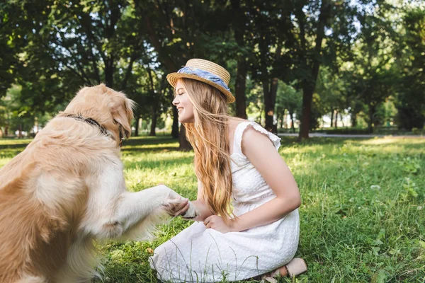 Full length view of beautiful girl in white dress and straw hat holding paw of golden retriever while sitting on meadow and looking at dog — Stock Photo