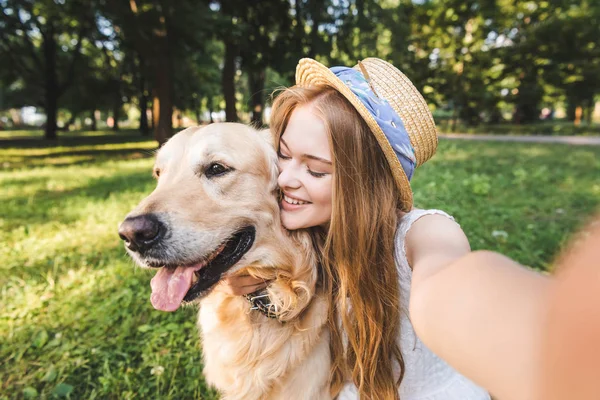 Young girl in white dress and straw hat hugging golden retriever and taking selfie while sitting on meadow and smiling with closed eyes — Stock Photo
