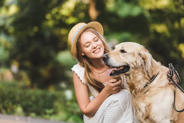 Hermosa chica en vestido blanco y paja sentado cerca de golden retriever y sonriendo mientras mira al perro - foto de stock