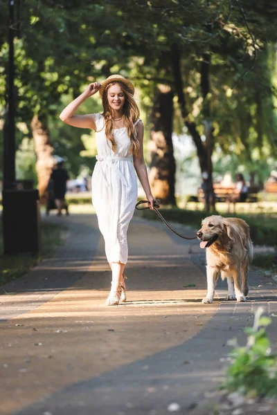 Full length view of beautiful girl in white dress and straw hat smiling while walking with golden retriever on pathway and looking away — Stock Photo