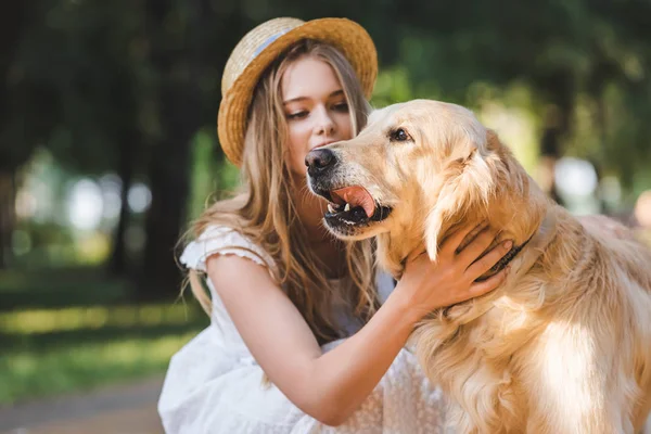 Schönes Mädchen in weißem Kleid und Stroh streichelt Golden Retriever und schaut Hund an — Stockfoto