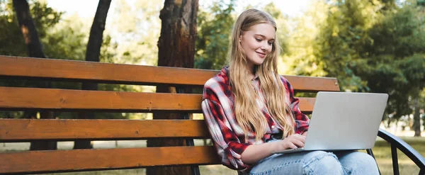 Tiro panorâmico de menina bonita em roupas casuais sentado no banco de madeira no parque e usando laptop — Fotografia de Stock