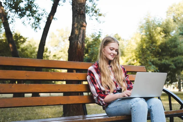 Beautiful girl in casual clothes sitting on wooden bench in park and using laptop — Stock Photo