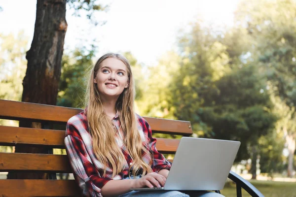 Bella ragazza in abiti casual seduto su una panchina di legno nel parco, utilizzando il computer portatile e guardando altrove — Foto stock