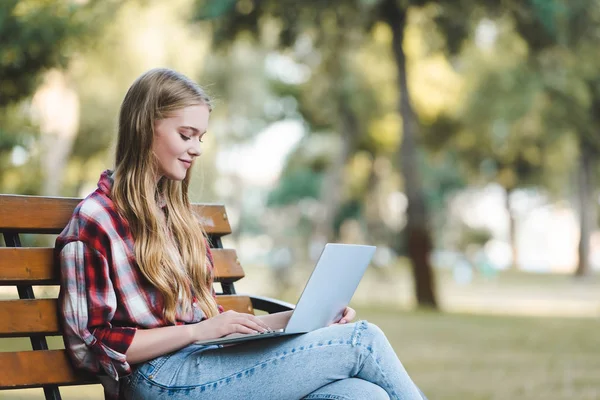 Selektiver Fokus der schönen Mädchen in lässiger Kleidung, die auf einer Holzbank im Park sitzen und Laptop benutzen — Stockfoto