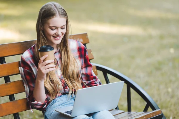 Hermosa chica en ropa casual sosteniendo taza de café de papel mientras está sentado en el banco de madera en el parque y el uso de la computadora portátil - foto de stock