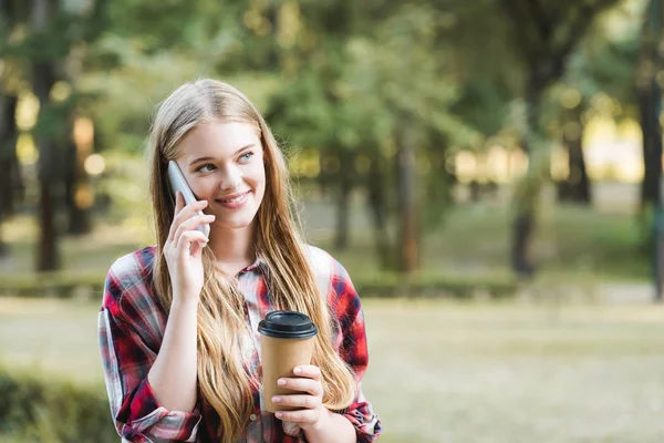 Messa a fuoco selettiva di bella ragazza in abiti casual in possesso di tazza di caffè di carta mentre si parla su smartphone e guardando altrove — Foto stock