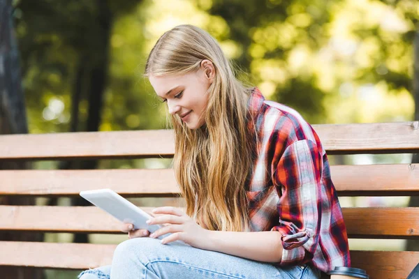 Foco seletivo da menina bonita em roupas casuais sentado no banco de madeira no parque e usando tablet digital — Fotografia de Stock