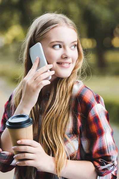 Retrato de la hermosa chica en ropa casual sosteniendo taza de café de papel y sonriendo mientras habla en el teléfono inteligente y mirando hacia otro lado - foto de stock