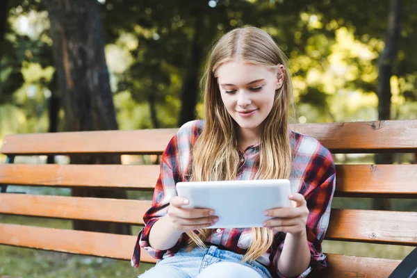 Chica joven en ropa casual sentado en el banco de madera en el parque y el uso de la tableta digital - foto de stock