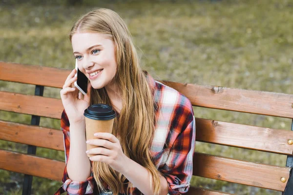 Belle fille en vêtements décontractés tenant tasse de café en papier tout en étant assis sur un banc en bois dans le parc et parler sur smartphone — Photo de stock