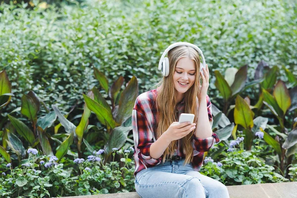 Hermosa chica en ropa casual sonriendo mientras escucha musicar en los auriculares - foto de stock